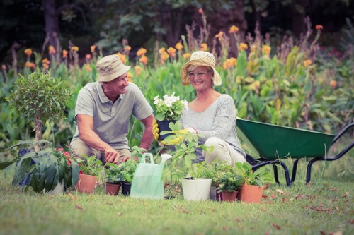A middle-aged couple in a lush, well-maintained backyard garden sit on the grass surrounded by an array of potted plants and flowering shrubs, with vibrant purple and orange flowers blooming in the background. The woman, wearing a wide-brimmed straw hat and light-colored clothing, holds a white flowering plant in her hands while smiling at her partner. The man, also wearing a straw hat and casual attire, appears to be engaged in gardening activities. Nearby, there is a green wheelbarrow, a pink watering can, and additional pots of various sizes containing healthy green plants, indicating ongoing gardening or planting work. The lawn is neatly cut, with a mix of dense grass and soil patches, and the garden scene is illuminated by natural daylight under a partly cloudy sky, creating a peaceful outdoor environment typical for outdoor gardening and landscaping care in Tottenham. This setting showcases a landscaped yard that would benefit from professional gardening services provided by Landscapers Tottenham.