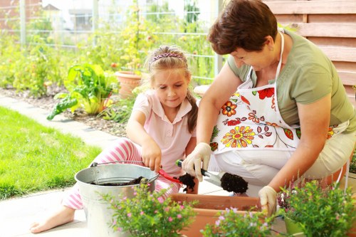 A woman and young girl are engaged in gardening activities in an outdoor backyard garden, with the woman wearing an apron and gloves while tending to a rectangular flower bed, and the girl assisting by planting or watering, both focused on the task. The garden features lush green grass, a variety of potted plants, and a mix of flowering and leafy shrubs along the fence in the background, with a wooden patio or deck area nearby. The scene is illuminated by natural daylight, suggesting a pleasant weather day in Tottenham, with a well-maintained outdoor space that highlights typical elements of suburban garden landscaping and outdoor maintenance suitable for professional gardening services.