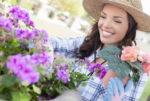 A woman wearing a straw hat and blue gloves is tending to a garden with various flowering plants, including purple and pink blooms, in a landscaped outdoor space. The garden features a well-maintained lawn, edged with flower beds and lush green foliage. In the background, there are trees and a paved patio area, suggesting a residential garden in Tottenham. The scene is brightly lit, indicating a sunny day, with natural tones of green, purple, and pink capturing the vibrant and healthy appearance of the plants. The image emphasizes outdoor gardening activities and landscaping care, aligning with professional gardening services provided by Landscapers Tottenham, focusing on sustainable maintenance and garden enhancement in the local area.