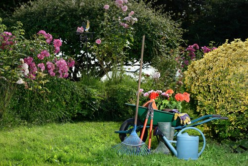 A neatly maintained garden with a vibrant green lawn in the foreground, bordered by a variety of flowering bushes and shrubs, including pink and purple blossoms and lush yellow-green foliage. In the middle ground, there are garden tools and equipment such as a blue watering can, a rake, a broom, and a wheelbarrow filled with gardening supplies placed on the grass. The background features tall trees and dense flowering plants creating a natural and lush outdoor environment. The scene suggests a well-kept outdoor space suitable for gardening tasks, with natural sunlight illuminating the garden area, typical of a residential garden in Tottenham. This setup reflects the expertise of landscapers offering garden maintenance and landscaping services in the local area, emphasizing the importance of outdoor care and landscaping improvements.