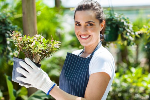 A young woman with dark hair tied back, wearing a white t-shirt, striped apron, and gardening gloves, stands in a lush garden. She is holding a small potted shrub with green and purple leaves, smiling at the camera. Behind her, there are vibrant green plants, trees, and foliage, with a blurred background suggesting a well-maintained outdoor space. The garden appears healthy and thriving, with dense plant growth and natural sunlight illuminating the scene, likely reflecting typical gardening activities associated with landscaping services in Tottenham. The setting emphasizes the professional approach to outdoor maintenance, lawn care, and horticultural enhancement provided by landscapers in the area, aligning with themes of gardening expertise and sustainable landscape management for residential or commercial gardens in the local postcode vicinity.