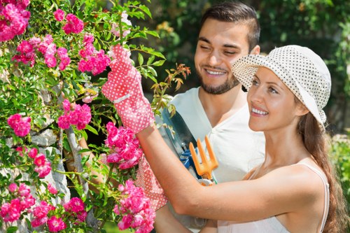 A woman and a man working together in a garden, with the woman pruning a vibrant pink flowering shrub using gardening gloves and holding a small hand tool, while the man stands nearby smiling. The garden features a lush green hedge, a well-maintained lawn with dense grass, and a paved pathway visible in the background, indicating an outdoor space in Tottenham or surrounding areas. Bright natural sunlight illuminates the scene, highlighting the greenery and flower colours, as they engage in garden maintenance activities typical of landscaping and gardening services provided by Landscapers Tottenham. The scene suggests late spring or summer weather, with the garden appearing tidy and cared for, reflecting professional outdoor maintenance.