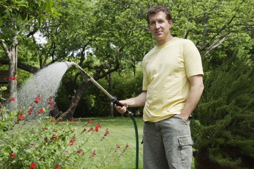 A man in a light yellow t-shirt and grey trousers stands in a well-maintained garden, holding a garden hose and watering a small flower bed with red and pink blooms. The garden features lush green grass, neatly trimmed bushes, and taller trees in the background, creating a natural and vibrant outdoor environment. The scene is outdoor under natural daylight, with a backdrop of leafy trees and dense foliage, indicating a healthy, flourishing green space. The garden layout includes a lawn area, flower beds, and landscaped shrubbery, typical of residential garden maintenance services in Tottenham or nearby areas. The man appears to be engaged in garden watering or maintenance, reflecting a focus on outdoor care and landscaping, consistent with the services offered by Landscapers Tottenham.