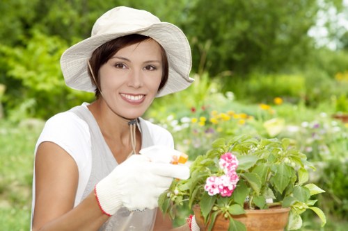 A woman wearing a wide-brimmed white garden hat, white gardening gloves, and a light-colored top is smiling while tending to a potted flowering plant with pink blossoms. She is in a lush outdoor garden environment with a variety of green foliage, including trees and shrubs, in the background. The garden features well-maintained flower beds, with visible soil and natural grass areas surrounding her. The scene is brightly lit, suggesting a sunny day, and the overall setting reflects a tidy, landscaped outdoor space suitable for gardening and lawn care services provided by landscapers in Tottenham. The photograph captures a moment of outdoor gardening activity within a carefully cared-for garden that emphasizes natural tones and healthy plant growth.