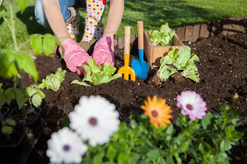 A person wearing pink gardening gloves is planting young lettuce plants in a well-maintained garden bed with dark, rich soil, surrounded by small gardening tools such as a yellow hand rake and a blue trowel. The garden features a variety of plants, with a focus on leafy greens in the centre, and other greenery visible in the background. Bright, colourful flowers including white, orange, and purple blooms are in the foreground, adding visual contrast and natural appeal. The garden appears to be part of a landscaped outdoor space, possibly a front or back garden, with a neatly edged border made of bricks or tiles. The environment is bathed in natural daylight, indicating a clear, sunny day, suitable for outdoor gardening activities. The photo conveys an active gardening scene, typical of garden maintenance and planting services provided by companies such as Landscapers Tottenham, supporting local garden renovation and upkeep in the Tottenham area. The overall setting combines natural floral colours with the lush green foliage and cultivated soil, emphasizing quality garden care and planting efforts to enhance outdoor spaces in London.