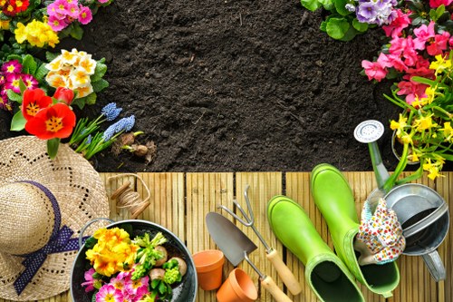 The image depicts a well-maintained garden border with dark, rich soil at the top, bordered by vibrant flower beds containing a variety of colorful flowering plants, including pink, yellow, and purple blooms. A narrow wooden decking area runs horizontally across the lower part of the scene, providing a clean, natural surface adjacent to the flower beds. On the decking, there are gardening tools such as a small trowel, a hand fork, and a pair of green rubber gardening boots, along with a decorative, wide-brimmed straw hat with a purple ribbon and a small pot filled with blooming flowers. The environment appears to be a tidy, landscaped backyard or front garden, suitable for outdoor gardening and maintenance activities, with natural daylight illuminating the scene. These elements highlight typical features of a garden ready for landscaping and lawn care services provided by Landscapers Tottenham, emphasizing the importance of garden layout, plant health, and outdoor aesthetic. The overall composition showcases a tidy, inviting outdoor space ideal for gardening work in the Tottenham area, linked subtly to local garden maintenance and landscaping services.