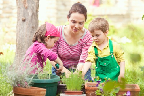 In a landscaped backyard garden with a lush green lawn, a woman with dark hair tied back is engaging with two young children—one girl wearing a pink headscarf and a pink top, and a boy in a yellow shirt with green overalls—while they work together tending to various potted plants and flower beds. The garden features a backdrop of mature trees and a garden fence, with the children reaching into pots containing herbs or small plants, and flower pots of different sizes and terracotta hues arranged on a garden table or surface. The scene captures a bright, outdoor environment under natural daylight, emphasizing the natural textures of the grass, soil, and wooden garden features, aligning with professional gardening and landscaping services offered by Landscapers Tottenham in the local area near the postcode TW, contributing to outdoor maintenance and sustainable gardening practices.