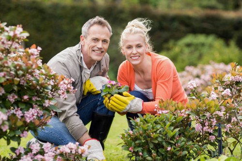 A man and a woman are gardening together in a lush, green outdoor space, smiling at the camera while kneeling among vibrant flowering shrubs with pink and white blossoms. The man is wearing a light grey shirt and yellow gardening gloves, holding a small potted plant or flower, while the woman is dressed in a coral-colored top, also wearing gardening gloves. The garden features neatly maintained flower beds with dense, healthy foliage, and a background of trees and hedges under natural daylight. This scene depicts outdoor gardening activities typical in a residential garden in Tottenham, with a focus on plant care and landscape maintenance that a professional gardening service such as Landscapers Tottenham could provide, including planting, pruning, and lawn care for a thriving, well-kept garden environment.